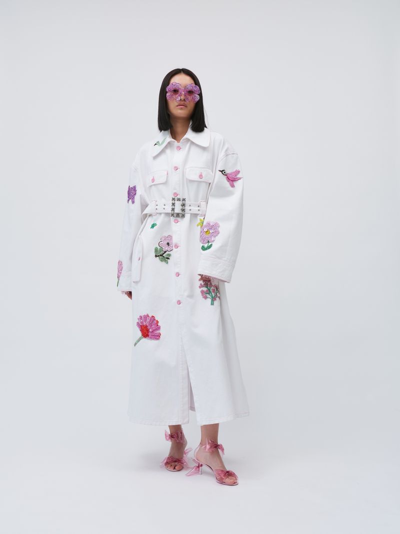 A woman in a white coat with flower embroidery and pink butterfly glasses poses in a photo studio with a plain white background.