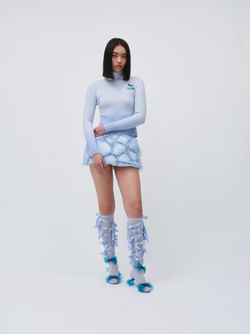 Full-length shot of a young Asian woman in a light blue gradient knit top and white and blue skirt, posing against a white backdrop.