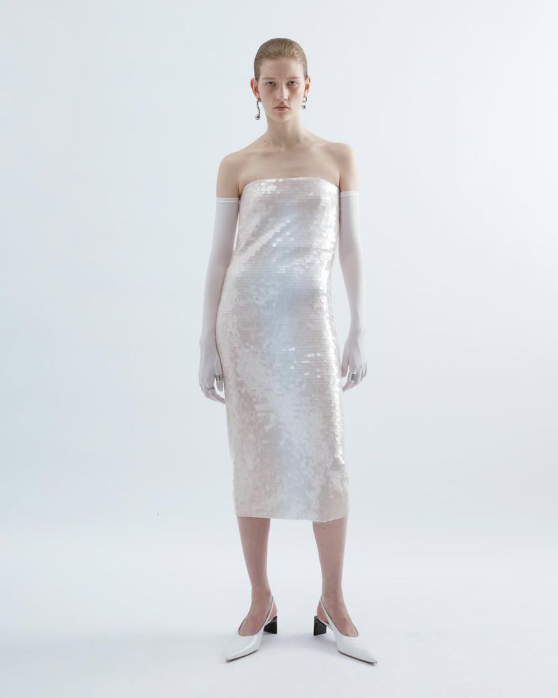 Elegant fair-skinned woman posing in a shimmering white strapless midi dress with long white gloves and white block-heeled pumps in a studio.
