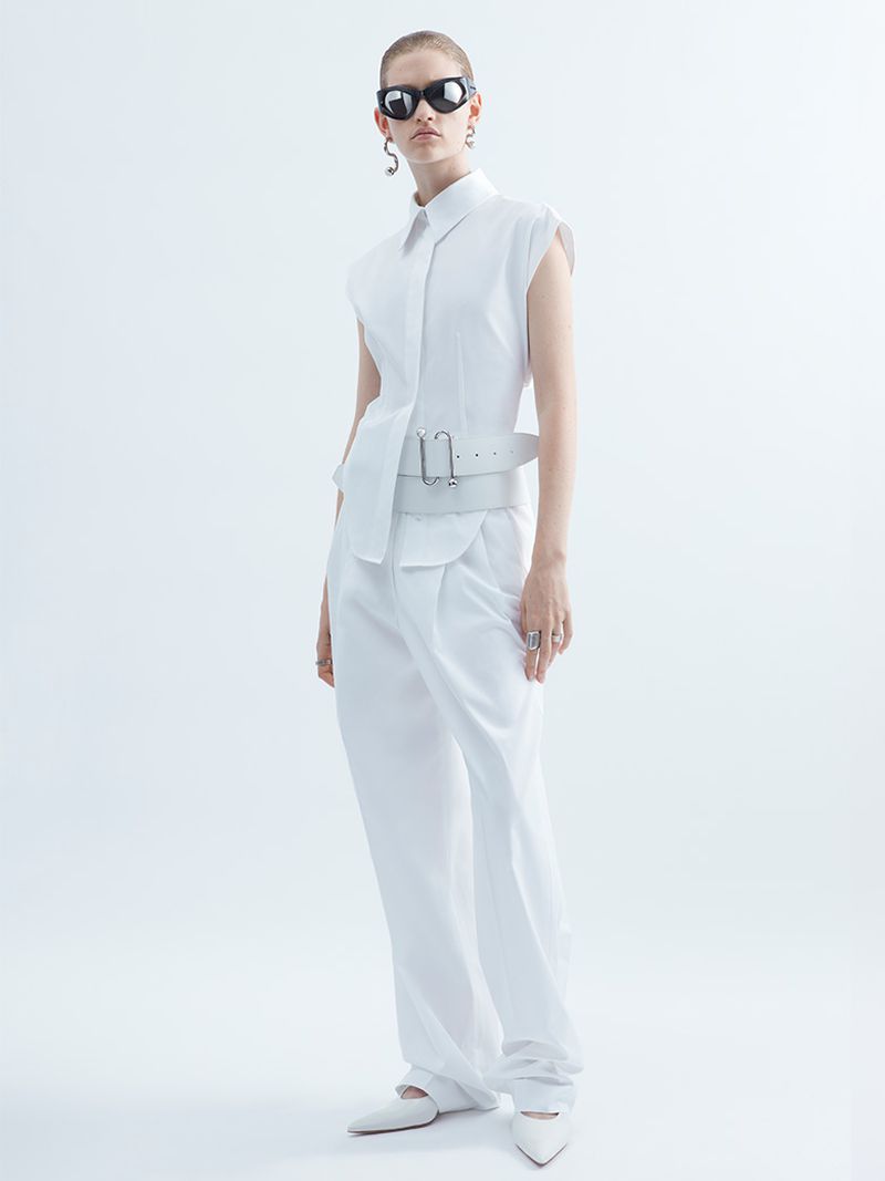 Fashion model in a white sleeveless shirt, wide-leg pants, and sunglasses poses in a bright, neutral studio setting for a fashion shoot.