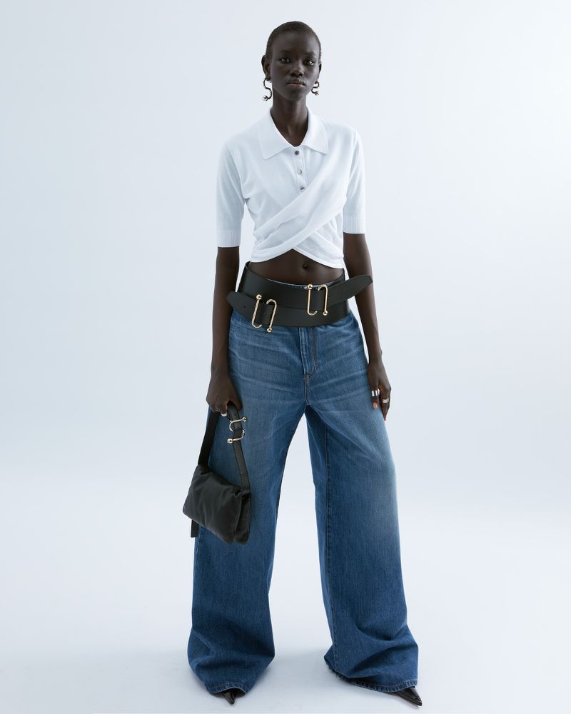 Studio shot of a black woman wearing a polo shirt, jeans, belt, and bag, standing against a white background with soft, diffused lighting.
