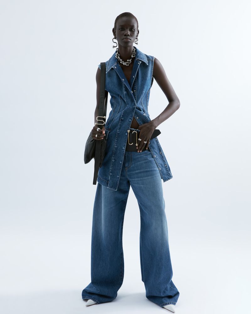 Fashion model poses in denim vest and wide-leg jeans against a white background, accessorized with a chain necklace and black shoulder bag.