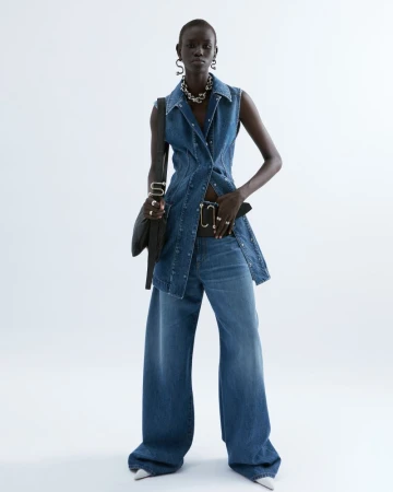 Fashion model poses in denim vest and wide-leg jeans against a white background, accessorized with a chain necklace and black shoulder bag.
