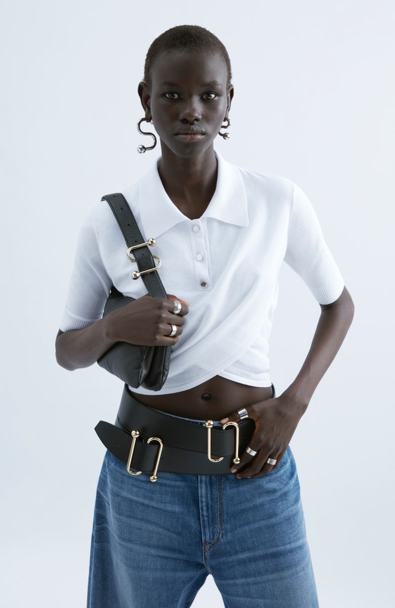 Fashionable dark-skinned woman wearing white polo top, blue jeans, leather belt, handbag, ring jewelry, studio lit on white background.