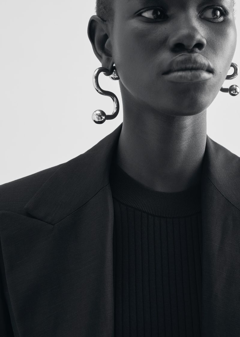 Close-up studio shot of a Black woman with silver earrings, wearing a black blazer and a black ribbed top against a white backdrop.