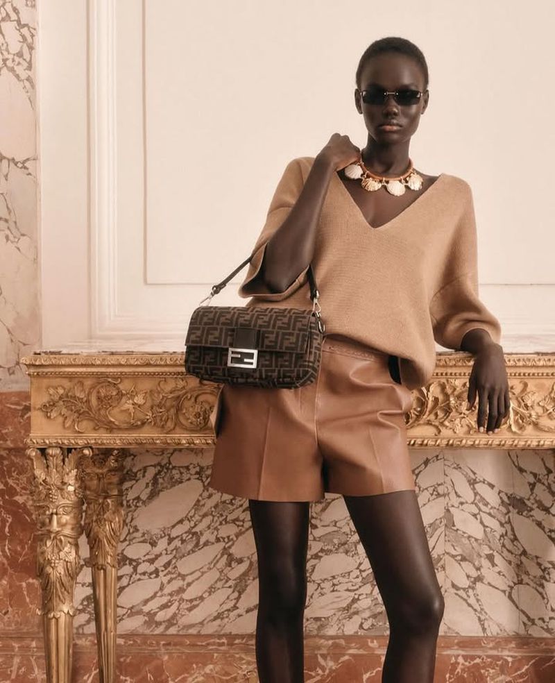 Fashion editorial shot of a Black woman wearing a tan sweater, leather shorts, necklace, sunglasses, and purse in an indoor setting.