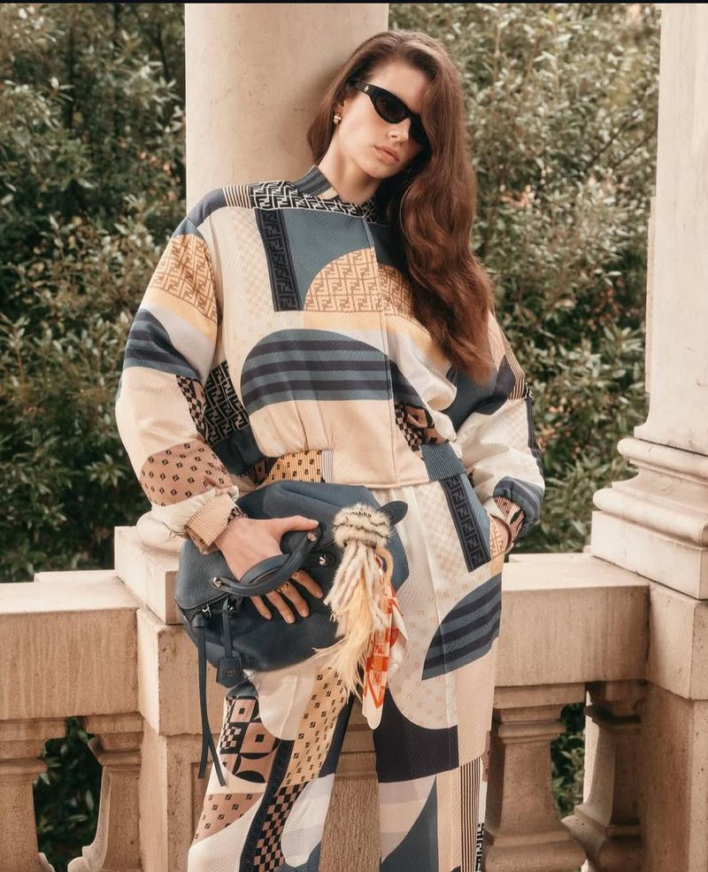 A woman with long brown hair wearing a patterned outfit, sunglasses, and carrying a black purse stands by a stone railing outdoors.