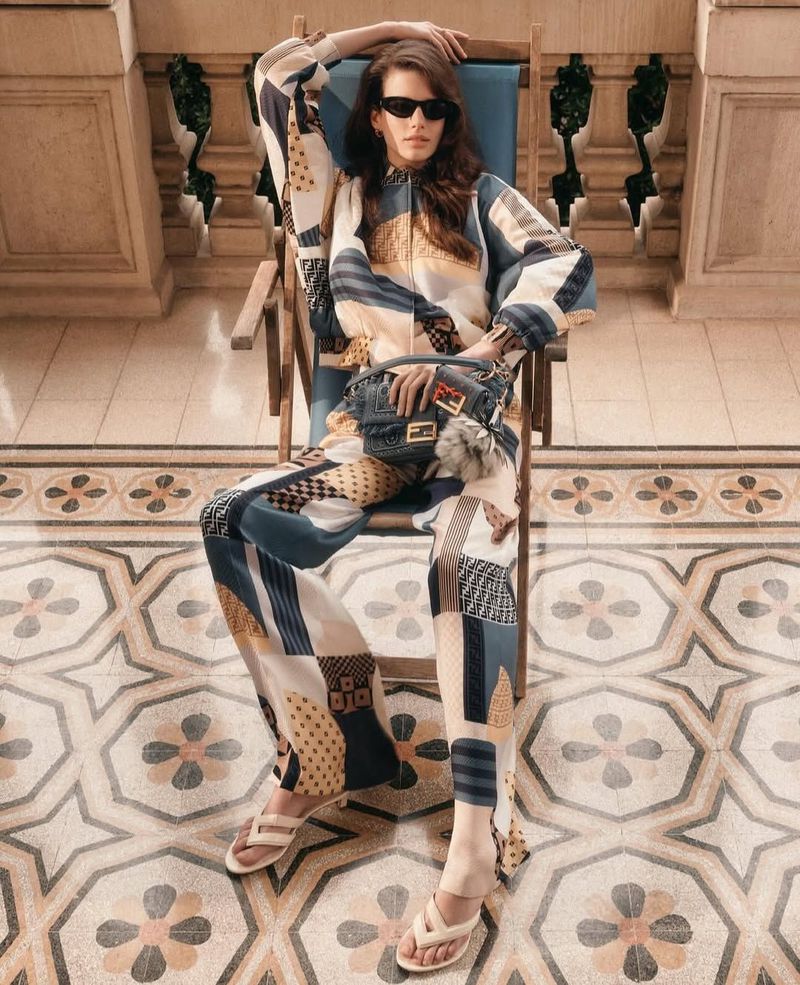 Adult female model sits in a chair wearing sunglasses, a patterned jacket and pants, and sandals. She is holding a small purse.