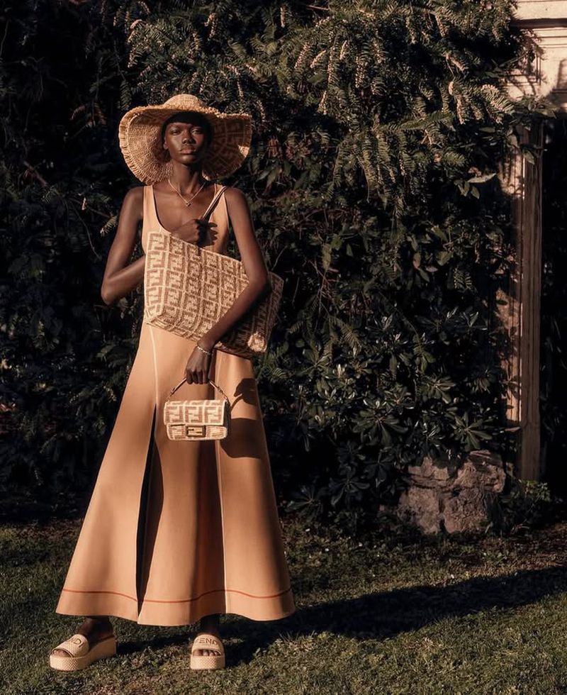 A tall, dark-skinned woman wearing a tan dress, straw hat, and sandals poses with two Fendi logo purses on a grassy lawn.
