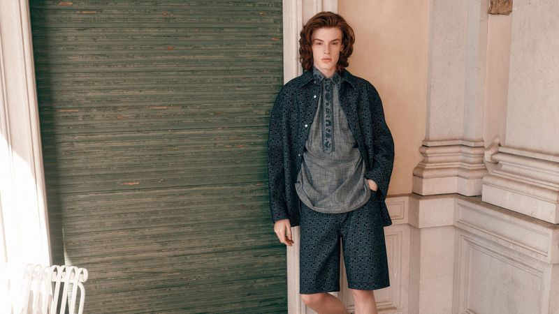 A red-haired male model poses in a patterned blue jacket, gray linen shirt, and matching patterned shorts in an indoor setting.