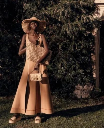 Model in a tan dress and wide-brim straw hat holding Fendi monogram bags outdoors.