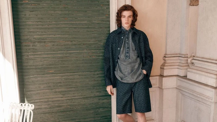A young male model with long reddish-brown hair wearing a dark patterned jacket, grey patterned shirt, and matching shorts standing outdoors