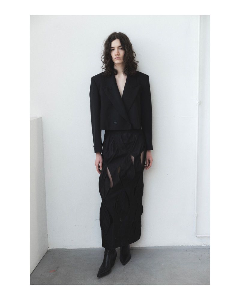 A slender adult model with long wavy hair poses indoors wearing a stylish black jacket, skirt, and boots against a white wall.