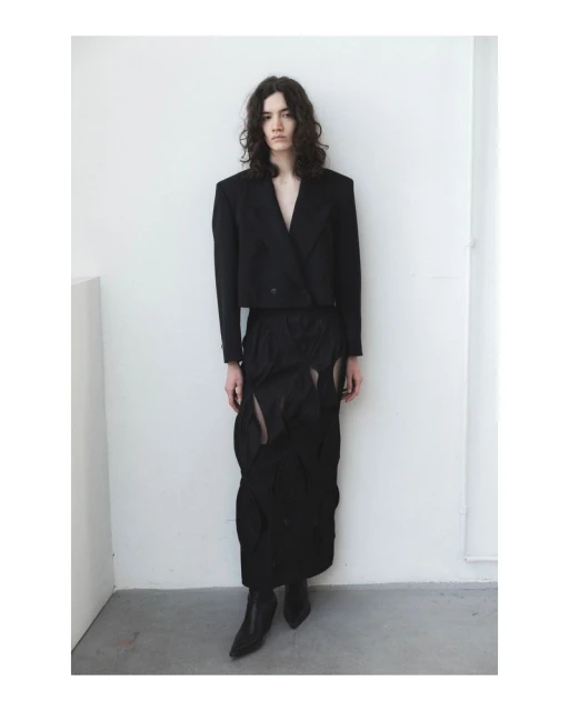 A slender adult model with long wavy hair poses indoors wearing a stylish black jacket, skirt, and boots against a white wall.