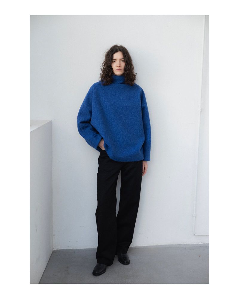 A woman with wavy brown hair poses indoors wearing a bright blue oversized sweater, black pants, and black shoes against a white wall.