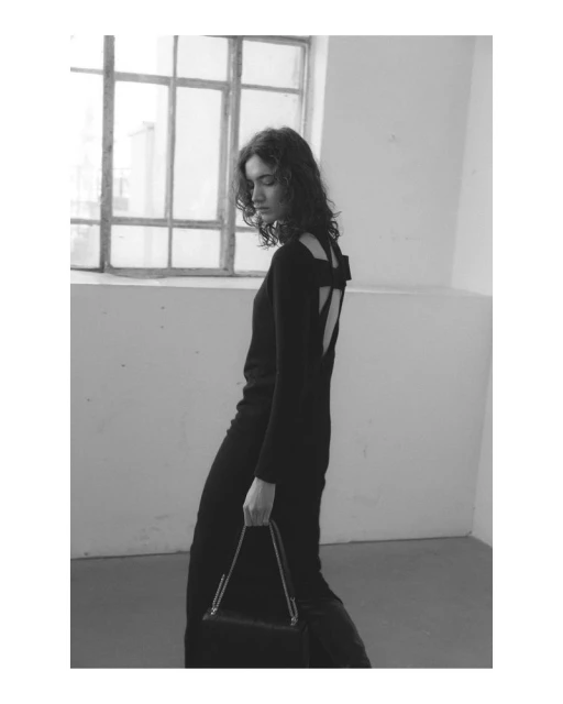 Elegant woman in a black dress with a back bow holding a purse with a chain in a minimalist indoor setting with natural light.