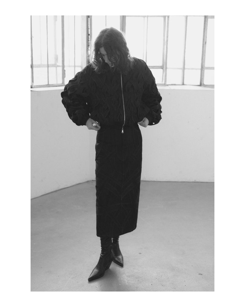 Monochrome studio shot of a stylish adult female in a black bomber jacket, pencil skirt, and ankle boots, standing against a window.