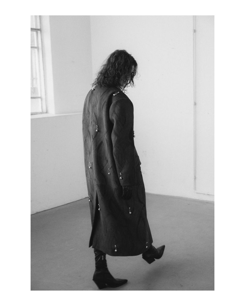 Fashionable woman in a long coat with pearl embellishments walks in a minimal white space, showcasing an editorial style.