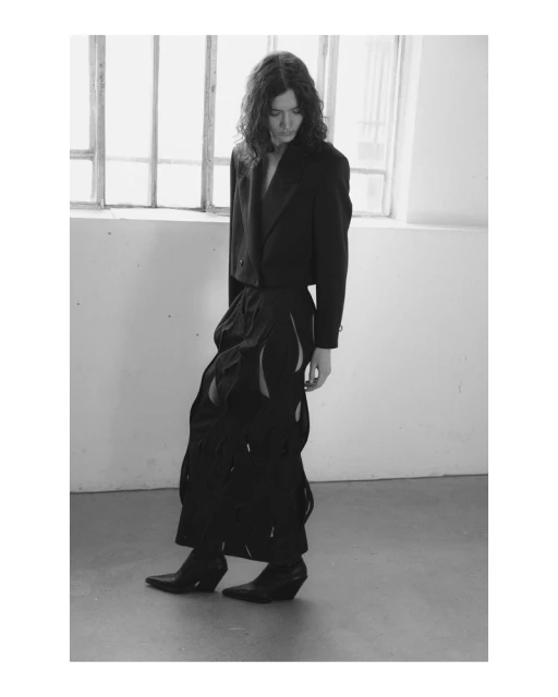 Black and white fashion shot of a slender adult standing indoors by a window wearing a blazer and pants with cut-out details and boots.