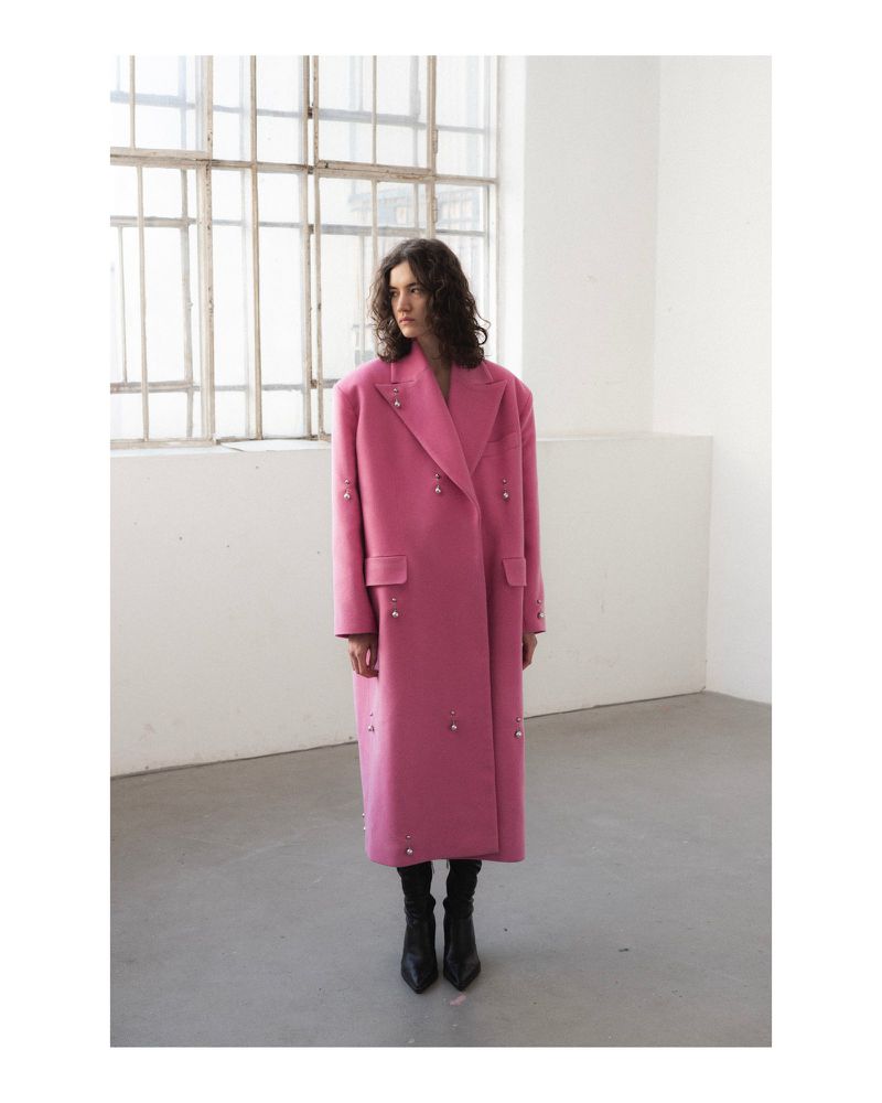 Woman in a pink overcoat with embellishments posing in a minimalist studio with large windows and natural light for a fashion editorial.