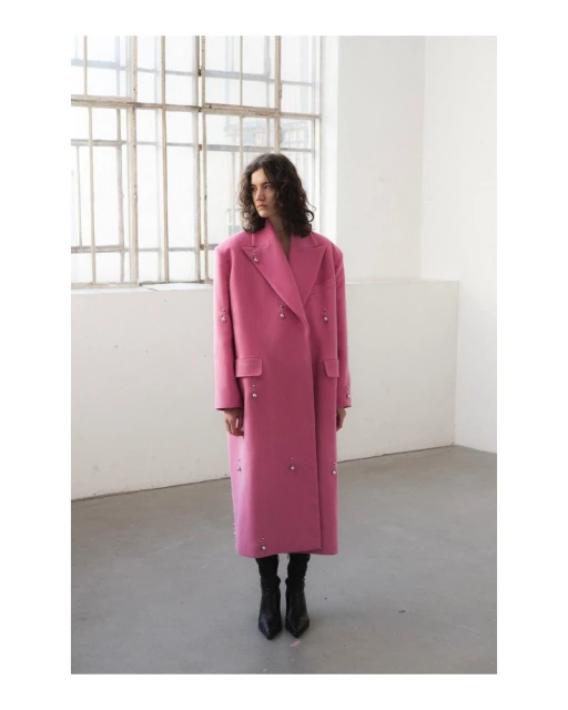 Woman in a pink overcoat with embellishments posing in a minimalist studio with large windows and natural light for a fashion editorial.