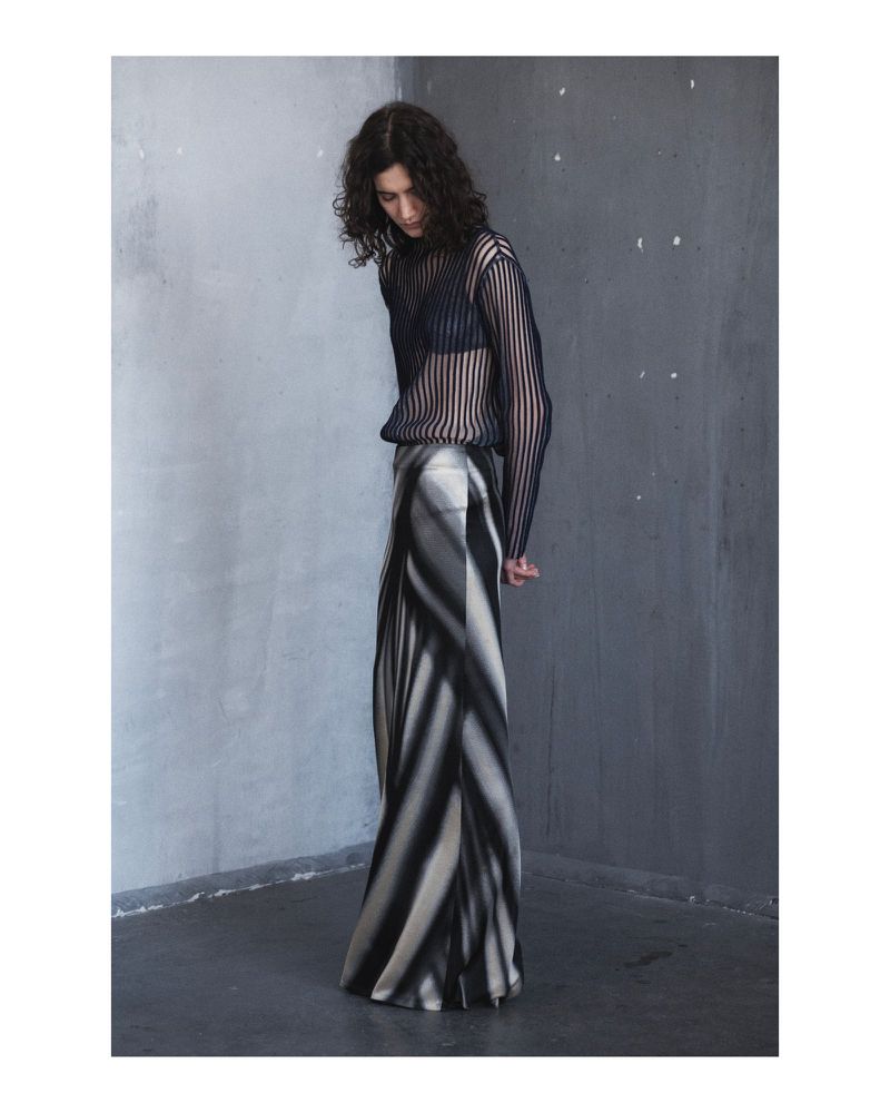 A woman with curly hair poses in a sheer black top and striped monochrome skirt against a mottled gray wall in a fashion shot.