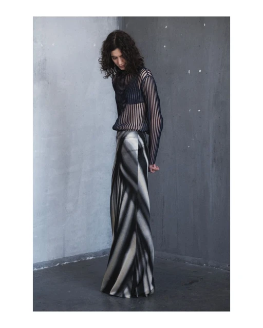 A woman with curly hair poses in a sheer black top and striped monochrome skirt against a mottled gray wall in a fashion shot.