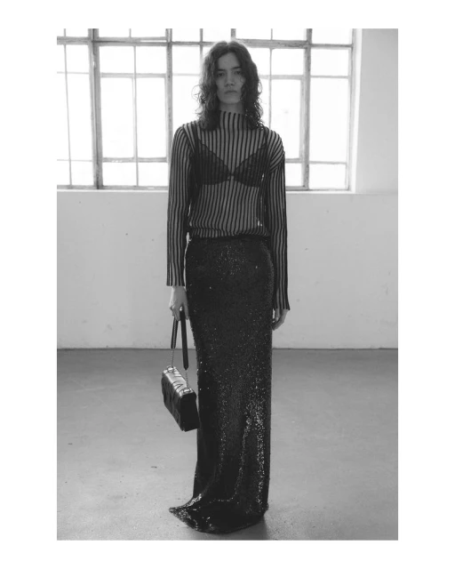 A woman stands in a striped top and black skirt holding a purse in a minimalist interior. Black and white, fashion shoot.