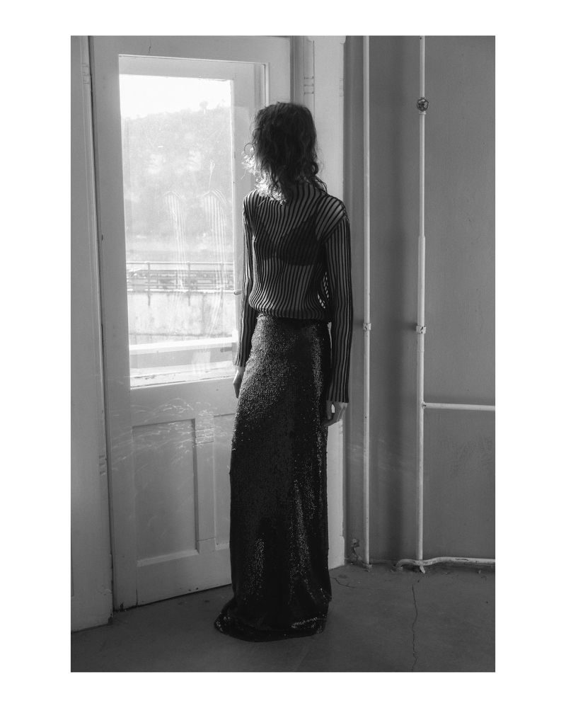 Back view of woman with curly hair in sheer top and sequin skirt standing by door in minimally furnished room, bright light.