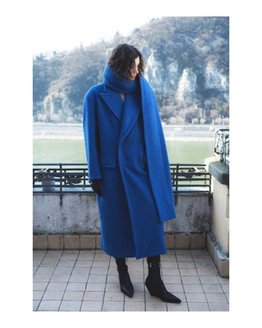 Woman in a striking blue overcoat and scarf stands on a tiled balcony with a mountain landscape and river backdrop on a sunny day.