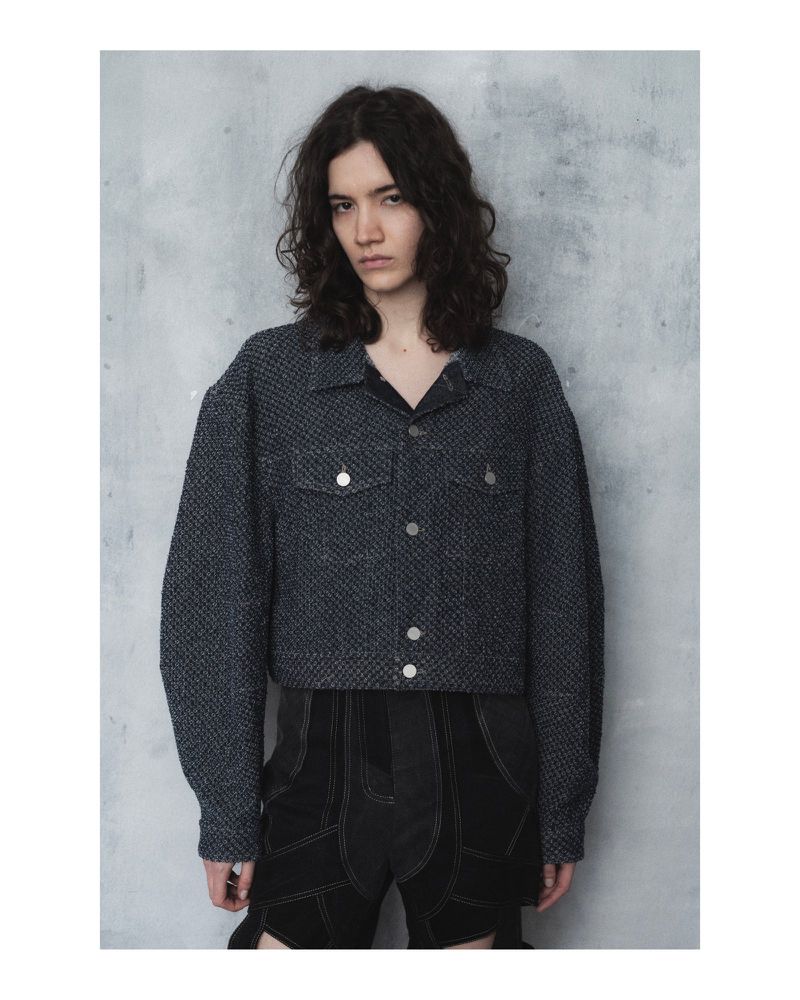 Fashionable person poses in a gray speckled jacket and dark shorts against a textured wall, capturing a contemporary, minimalist vibe.
