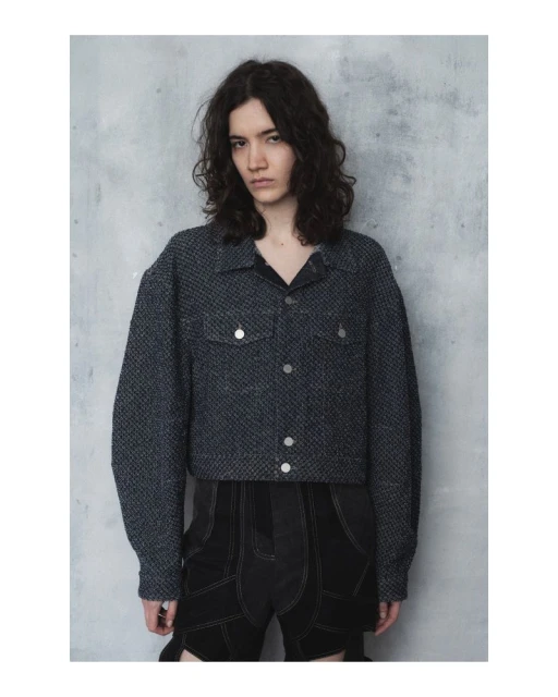 Fashionable person poses in a gray speckled jacket and dark shorts against a textured wall, capturing a contemporary, minimalist vibe.