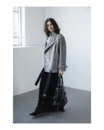 Model wearing a patterned grey jacket and black trousers holding a black handbag inside a bright studio