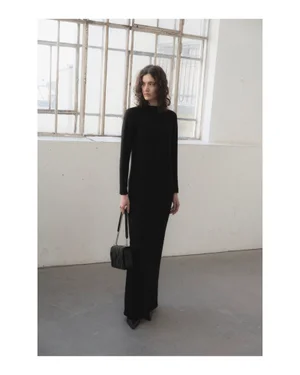 Model wearing a long black dress holding a black handbag standing indoors against large multi-paned windows.