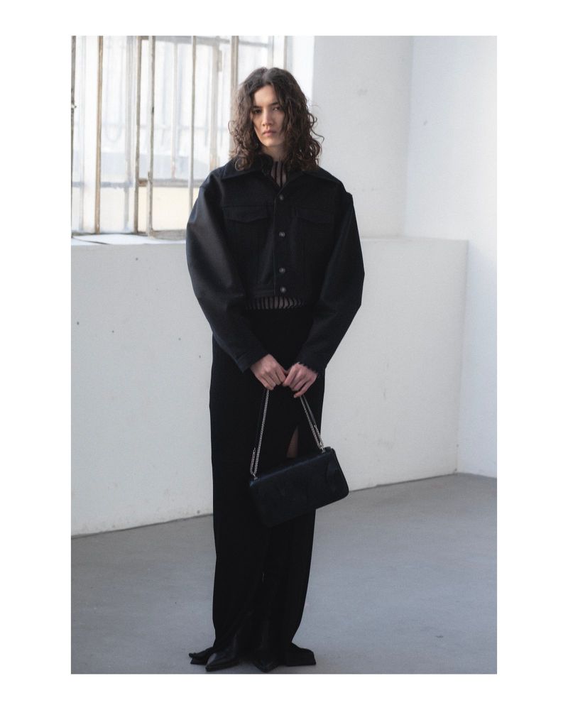 Model with long dark wavy hair wearing a black jacket and long skirt holding a black handbag inside an industrial room.