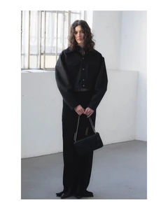 Model with long dark wavy hair wearing a black jacket and long skirt holding a black handbag inside an industrial room.