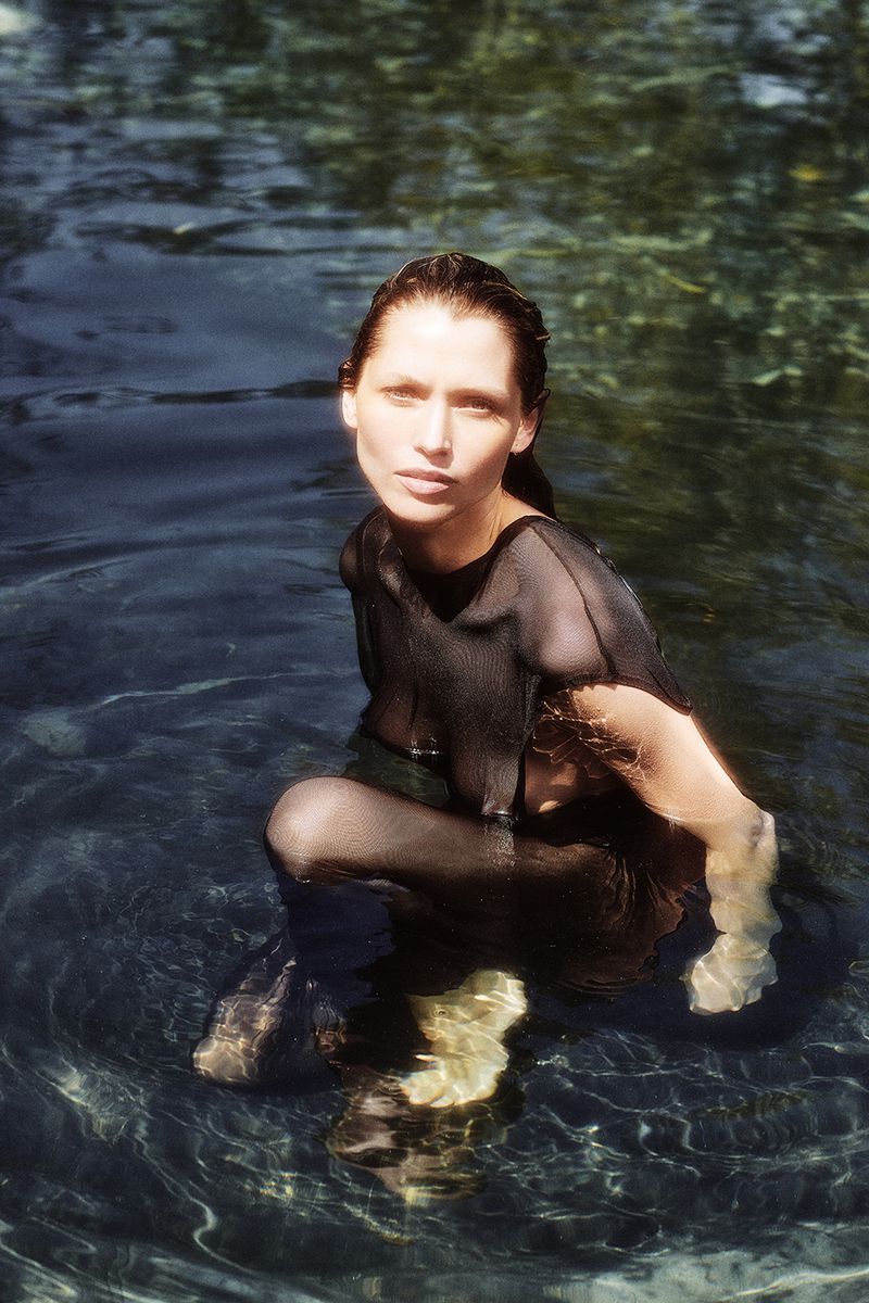 Woman in a black, semi-sheer shirt kneels waist-deep in water, her hair slicked back, set against a softly blurred background of trees and foliage.