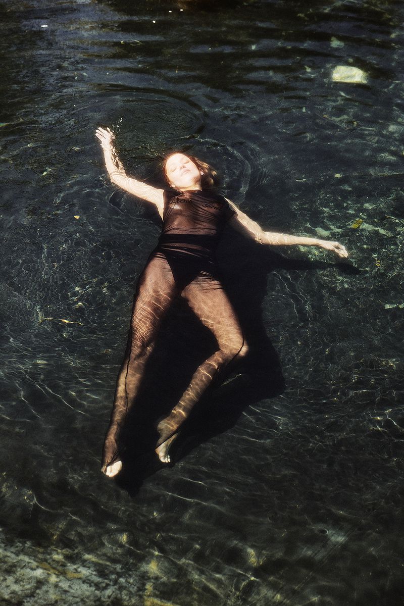 A woman in a black bodysuit floating in clear water, creating a serene scene with dappled light and reflections.