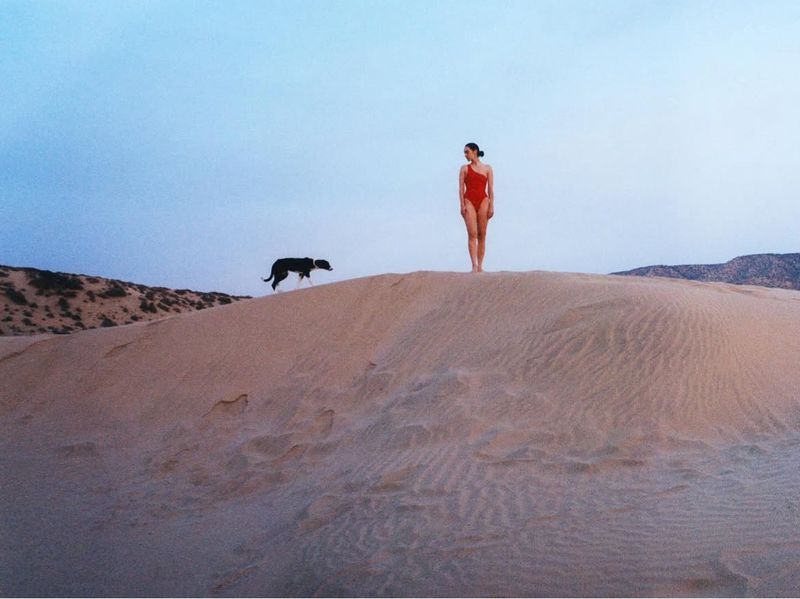Woman in red swimsuit poses on sand dune as black dog runs behind her against a bright blue sky at dusk near mountains.