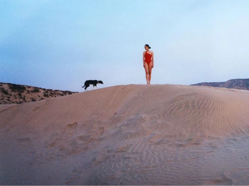 Woman in red swimsuit poses on sand dune as black dog runs behind her against a bright blue sky at dusk near mountains.