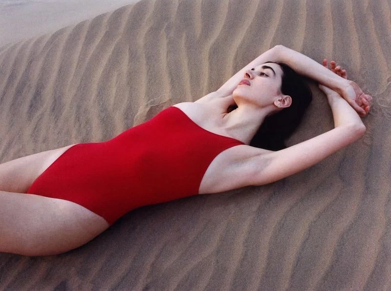Woman in red swimsuit lying on sand dunes with arms crossed above her head, illuminated by soft lighting in an outdoor shot.