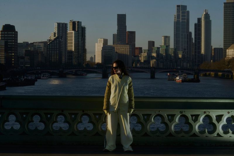 Person in casual outfit stands on bridge with cityscape, river, and boats in background, captured in a medium shot.