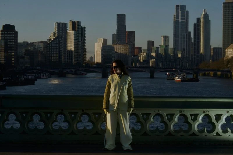 Person in casual outfit stands on bridge with cityscape, river, and boats in background, captured in a medium shot.