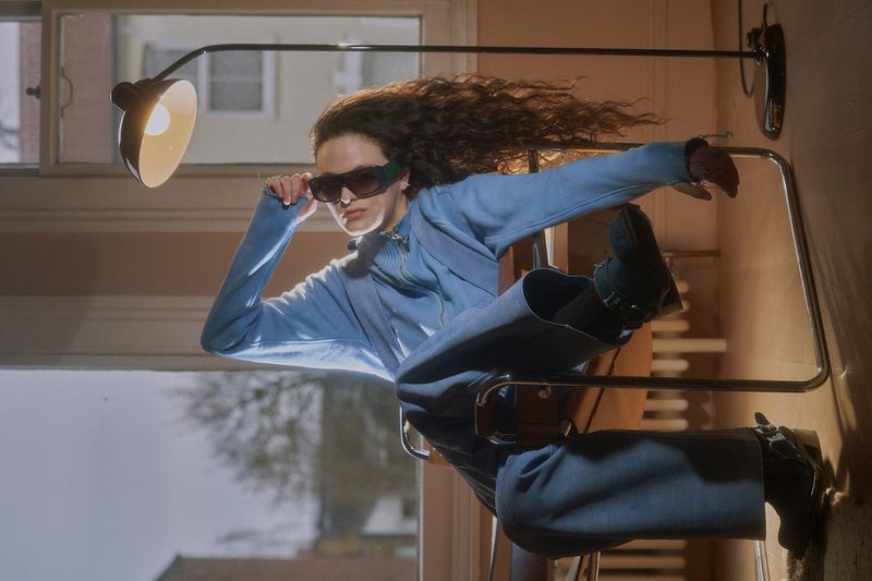 Young woman with curly hair poses in blue pantsuit and sunglasses in an indoor setting with natural light and a black adjustable lamp.