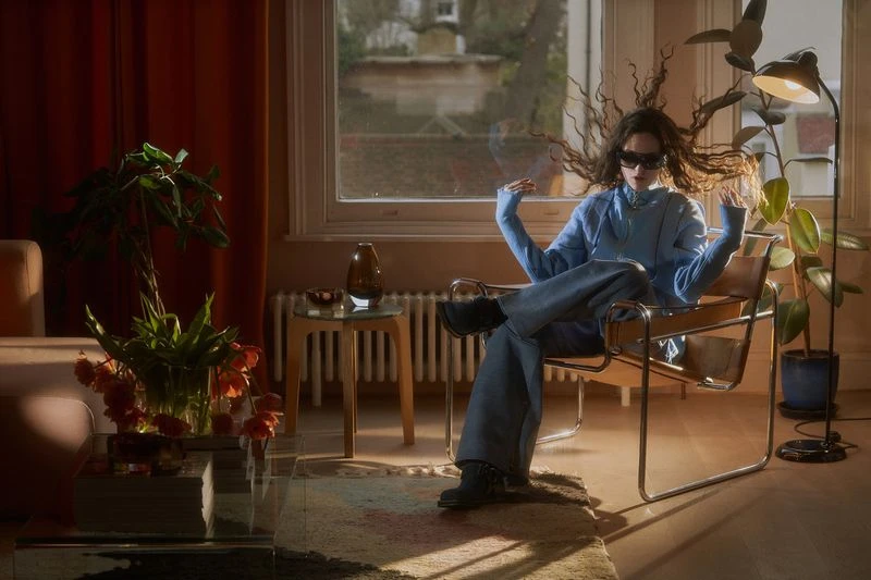 Woman in blue loungewear and sunglasses poses in modern chair indoors. Interior has plants, window, glass table, and warm tones.
