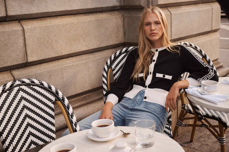 A blonde woman in a black and white cardigan sits at a cafe table with a coffee cup, a glass of water, and stone building in the background.