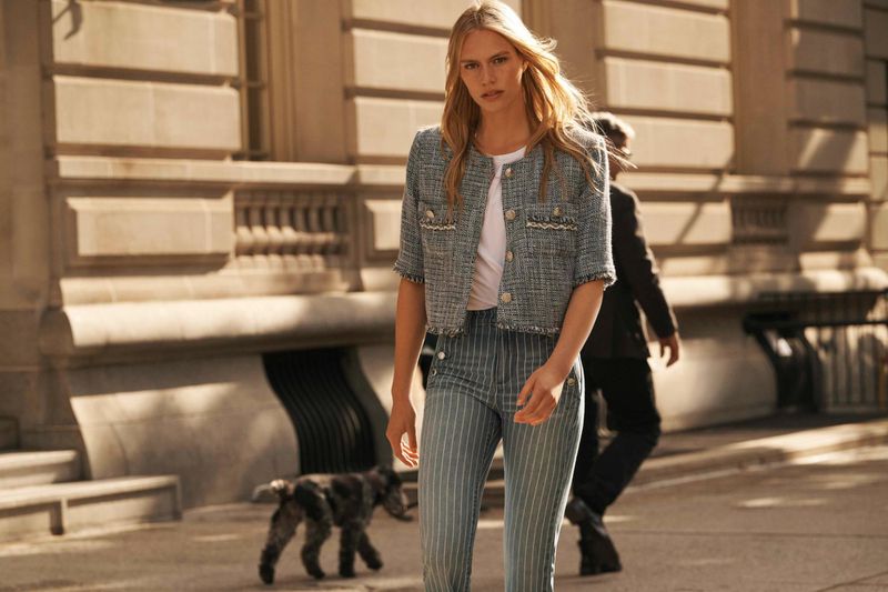 A blonde woman in a tweed jacket and pinstripe pants walks a dog on a city street with a man walking in the background.
