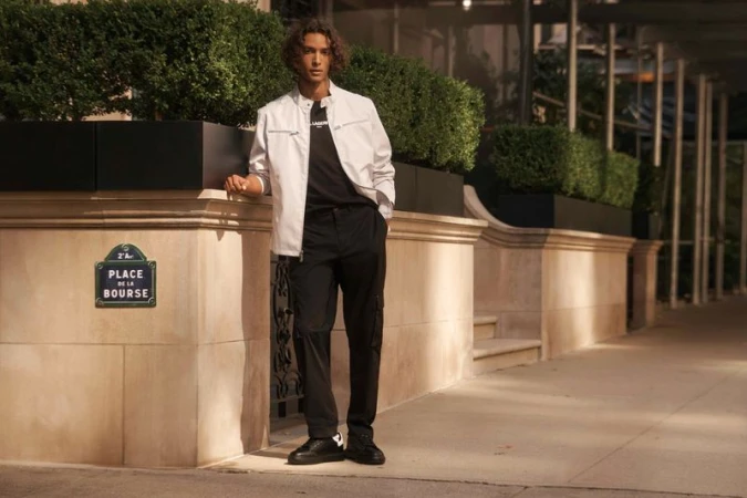 Man in white jacket and black pants standing in an urban setting with planters and a street sign in the background. Fashion.