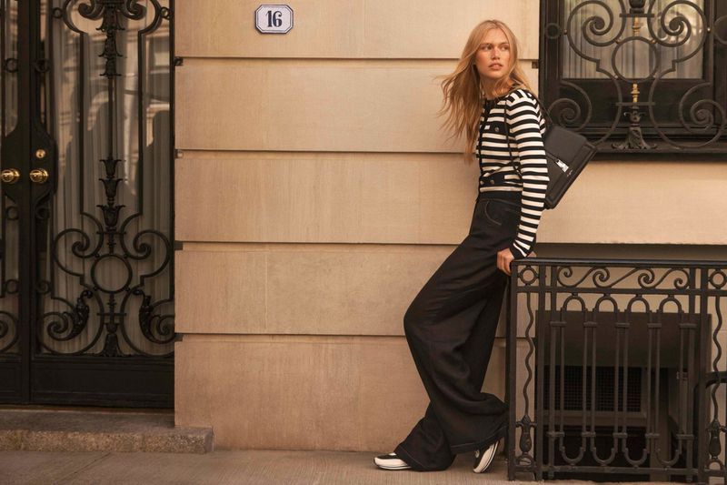 A blonde woman in a striped sweater and wide-leg pants poses against a building with an iron gate in an urban environment.