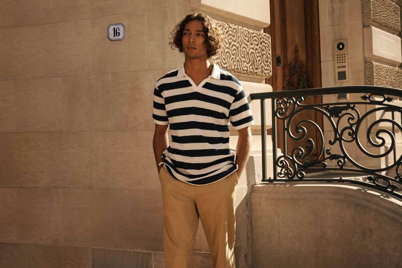 A young adult man in a striped polo shirt stands outside a building with a wrought-iron railing on a sunny day, looking away.